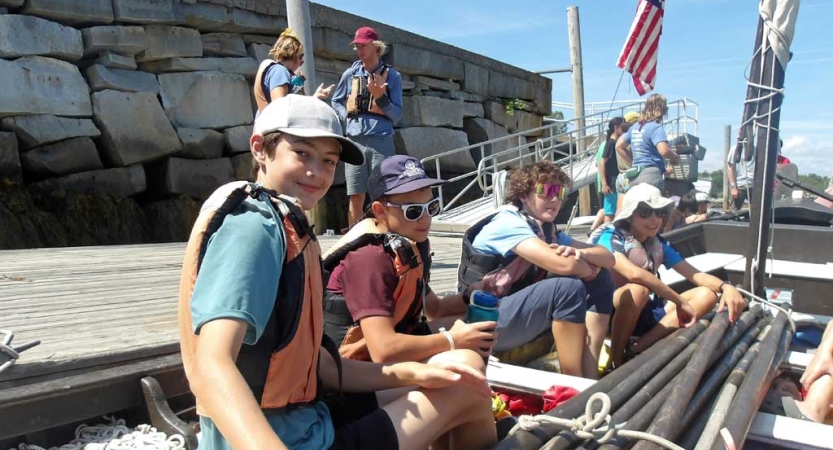 A group of students wearing life jackets sit in a boat that is docked. 